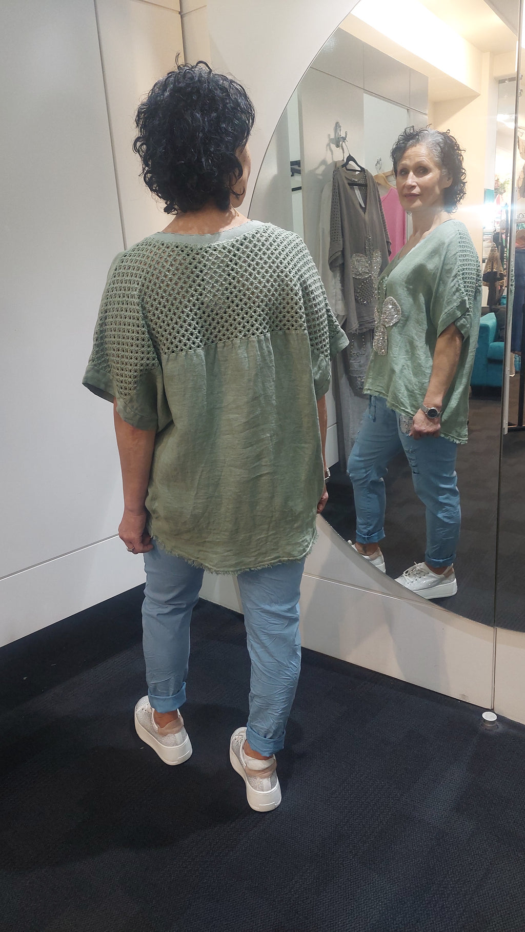 Woman wearing a green top with a butterfly design and light blue jeans, standing in front of a mirror.