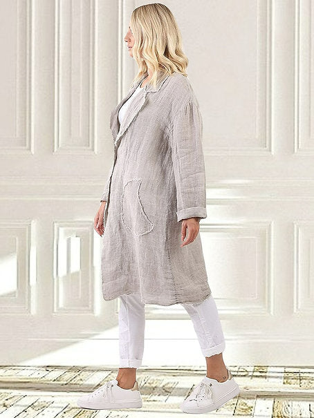 Woman wearing a long beige coat over a white outfit in front of a wooden wall.