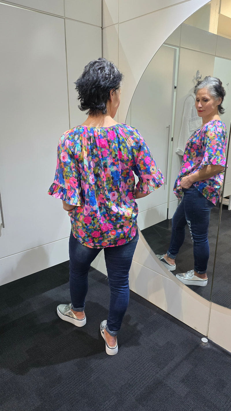 Person wearing a colorful floral blouse standing in front of a mirror. Tie Neck Top Multi