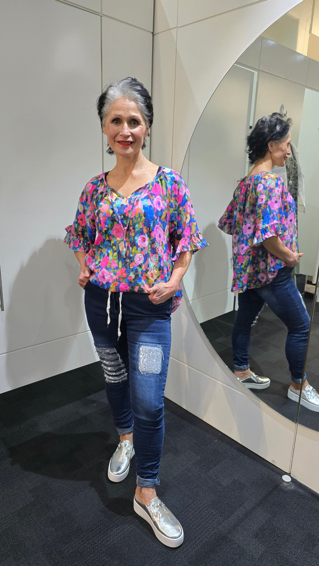 Woman wearing a colorful floral blouse and jeans standing in front of a mirror. Tie Neck Top Multi