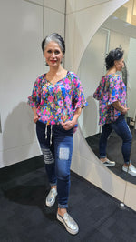 Woman wearing a colorful floral blouse and jeans standing in front of a mirror. Tie Neck Top Multi