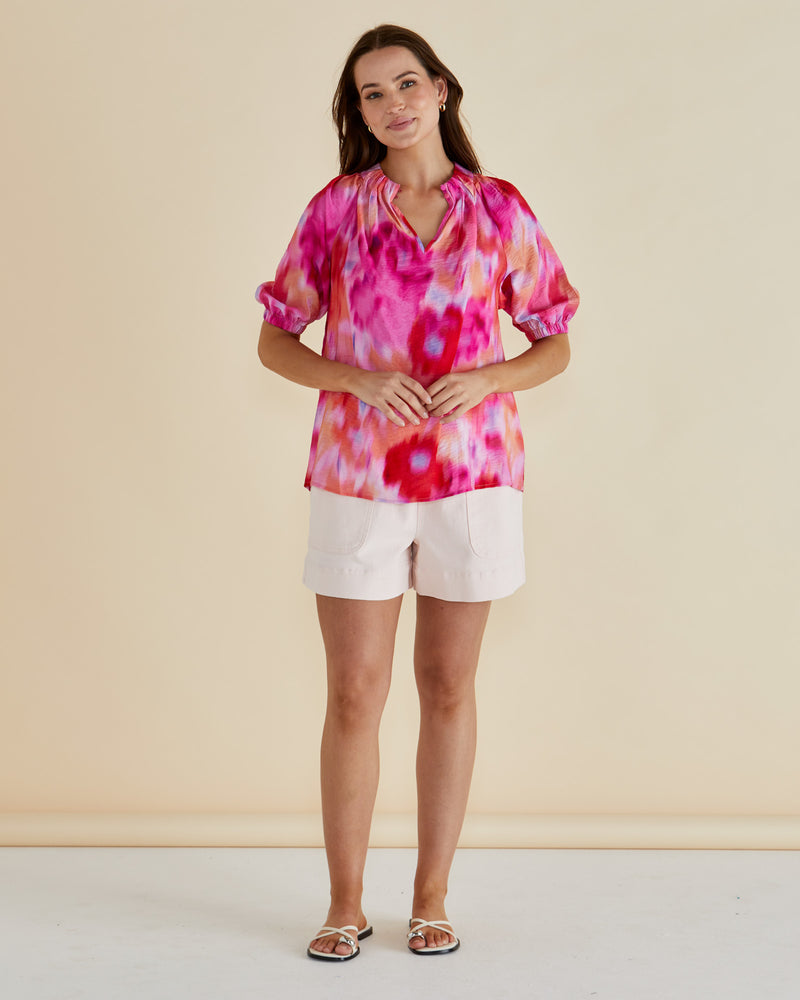 Woman wearing a colorful tie-dye top and white shorts on a beige Joanne Blouse Floral Pink background
