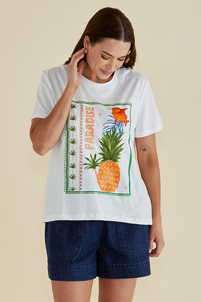 Woman wearing a white t-shirt with a pineapple graphic and 'Paradise' text on a beige background Ellia Crew Tee White
