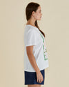 Woman wearing a white t-shirt with a pineapple graphic and 'Paradise' text on a beige background Ellia Crew Tee White