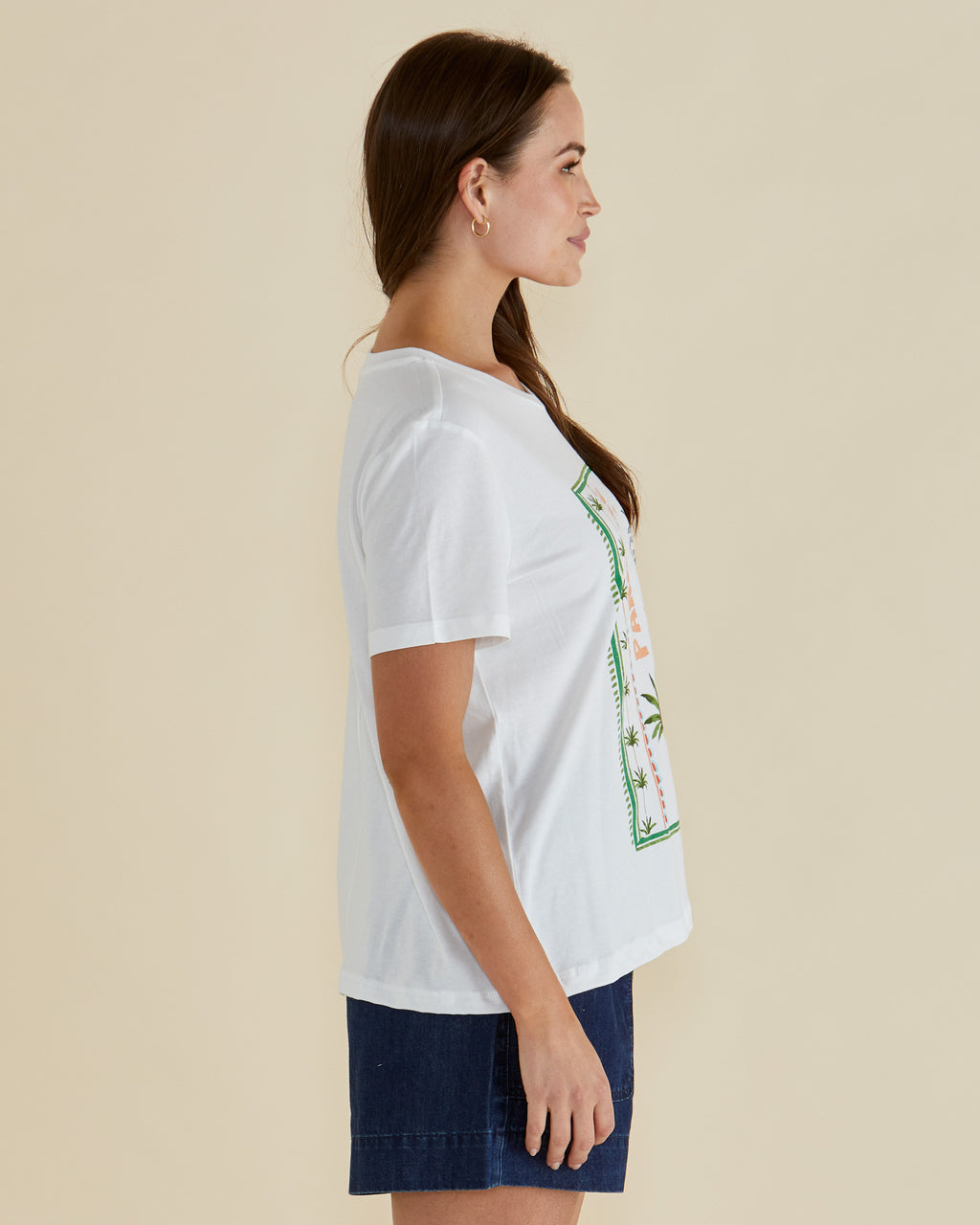 Woman wearing a white t-shirt with a pineapple graphic and 'Paradise' text on a beige background Ellia Crew Tee White