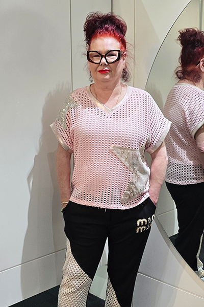Person wearing a light pink knit top with floral patterns and black pants, standing against a white wall. 
Italian Metallic Stars Top Pink