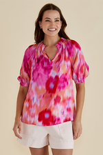 Woman wearing a colorful tie-dye top and white shorts on a beige Joanne Blouse Floral Pink background