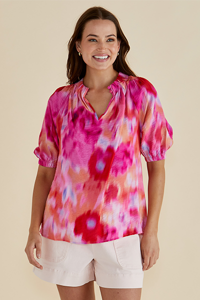 Woman wearing a colorful tie-dye top and white shorts on a beige Joanne Blouse Floral Pink background