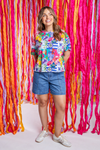 Woman wearing a colorful patterned shirt and denim shorts standing in front of a vibrant, multicolored ribbon background.
Lala Land Tee