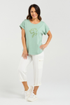 Woman wearing a light green t-shirt with a graphic design on a white background Lily Top Sage