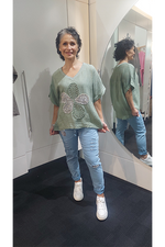 Woman wearing a green top with a butterfly design and light blue jeans, standing in front of a mirror.
