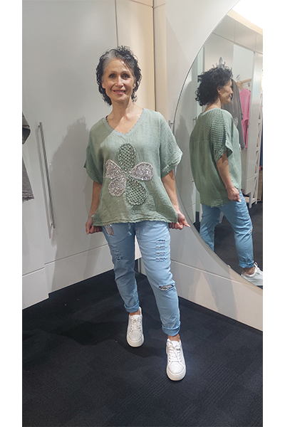 Woman wearing a green top with a butterfly design and light blue jeans, standing in front of a mirror.