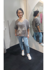 Woman standing in a fitting room wearing a Taupe t-shirt with floral design and blue jeans.