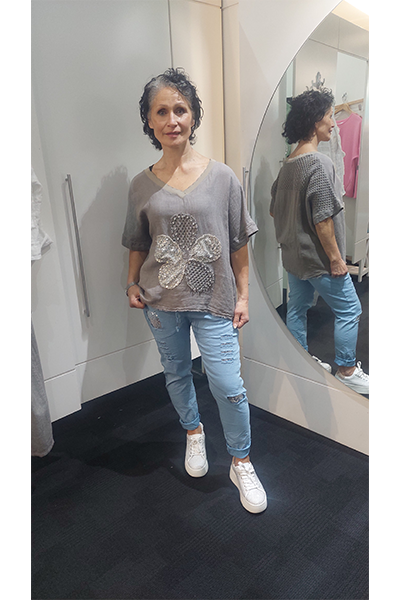 Woman standing in a fitting room wearing a Taupe t-shirt with floral design and blue jeans.
