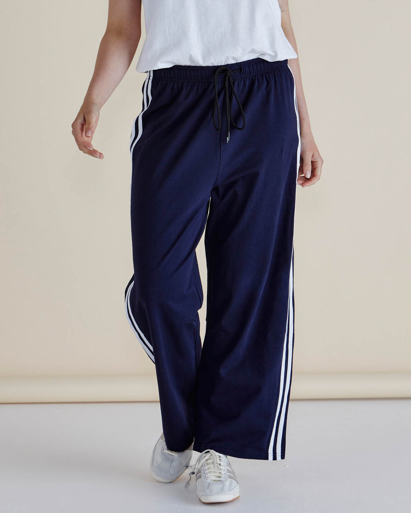 Navy track pants with white stripes worn by a person on a beige background