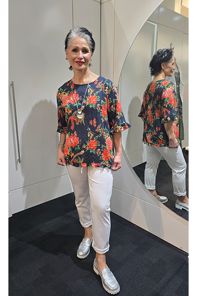 Woman wearing a floral blouse and white pants standing in front of a mirror. Round-Neck-Top-Multi