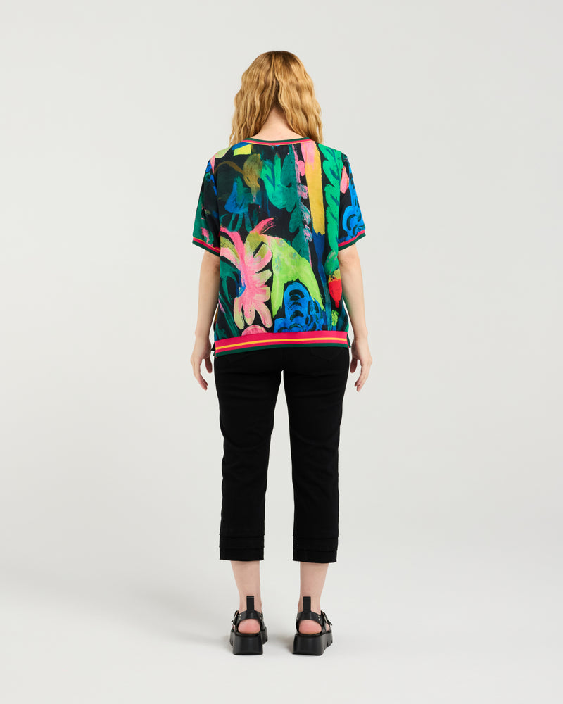 Person wearing a colorful tropical-themed shirt and black pants on a white background Zoe Top Midnight Fern