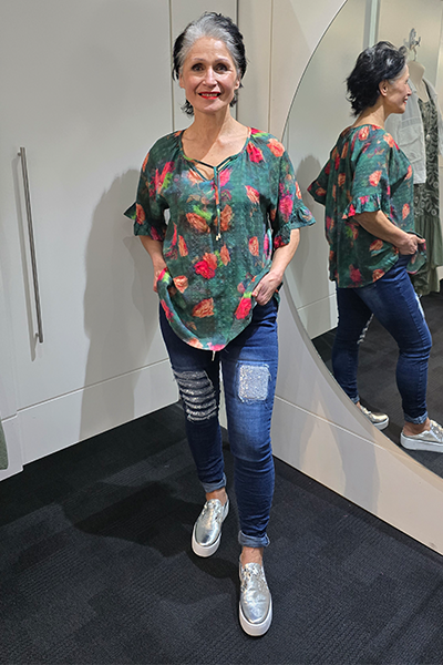 Woman standing in front of a mirror wearing a colorful shirt and blue jeans. Tie Neck Top Green