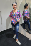 Woman wearing a colorful floral blouse and blue jeans standing in front of a mirror. Tie Neck Top Multi