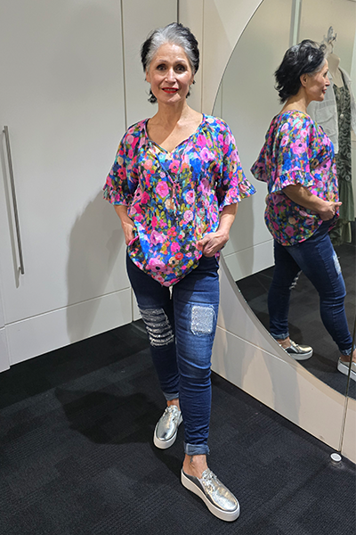 Woman wearing a colorful floral blouse and blue jeans standing in front of a mirror. Tie Neck Top Multi