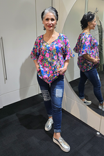 Woman wearing a colorful floral blouse and blue jeans standing in front of a mirror. Tie Neck Top Multi