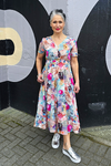Woman in a floral dress standing against a black wall with large white letters.
V Neck Dress Multi