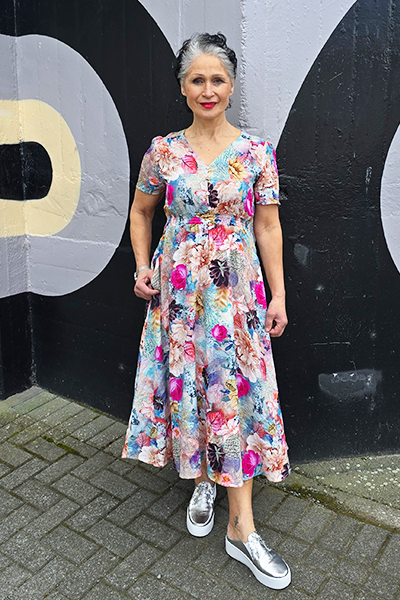 Woman in a floral dress standing against a black wall with large white letters.
V Neck Dress Multi