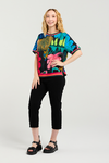 Woman wearing a colorful floral-patterned shirt and black pants on a white background Zoe Top Midnight Fern