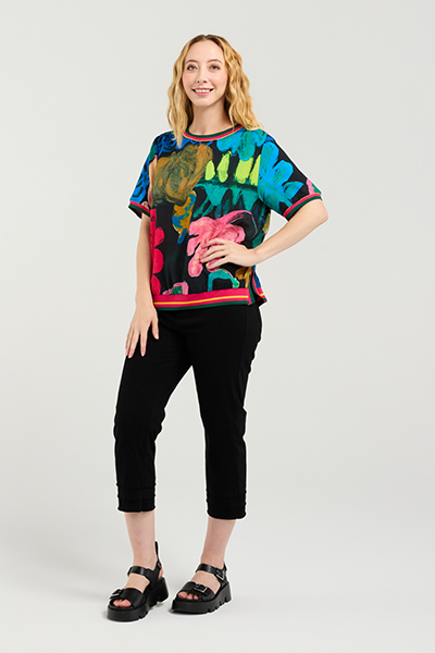 Woman wearing a colorful floral-patterned shirt and black pants on a white background Zoe Top Midnight Fern